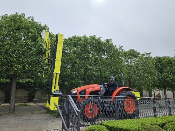 A red tractor with a tall yellow shearing device that is being used to trim trees. 